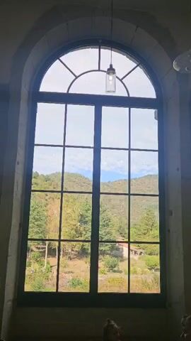 The video shows a large arched window with multiple panes. Through the window, a landscape with rolling hills covered in trees and vegetation is visible. The sky is partly cloudy. In the foreground, out of focus, the top of two heads of people can be seen. A single light fixture hangs from the ceiling above the window. The window frame appears to be made of dark wood or metal, and the surrounding wall is stone. The overall impression is of an indoor space looking out onto a natural scene.