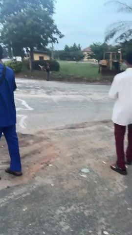 The video shows a black Toyota Tacoma pickup truck driving on a paved road, passing in front of three people who are standing on a dirt and gravel surface. One person is holding a small, light-colored object, possibly a piece of cloth or paper, while the other two people are wearing blue and maroon colored clothes respectively. There is a building, trees, and vegetation on the background. The pavement shows patches of repair and dirt. The video is shot outdoors in daylight.