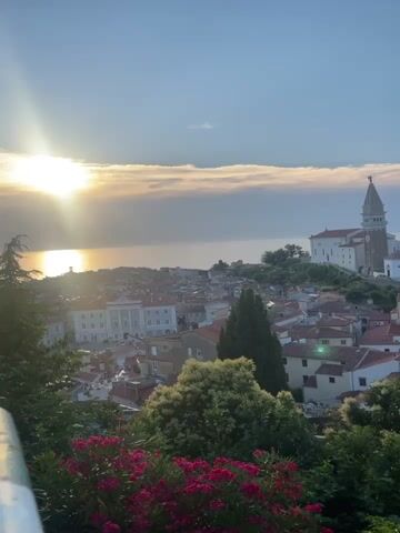 A high-angle, handheld video shows a scenic view of a coastal European town during a sunset. The sun, a bright orb with lens flares, is positioned in the upper left quadrant of the frame, partially obscured by wispy clouds. The sea stretches out towards the horizon, reflecting the warm hues of the setting sun. The town itself is a dense collection of buildings with red-tiled roofs, nestled on a hill. Dominating the skyline on the right is a tall, white church with a bell tower topped with a cross. Lush green trees and vibrant pink flowers fill the foreground, creating a natural frame for the picturesque landscape. The sky is a clear blue, with a few scattered clouds adding texture. The overall impression is one of tranquility and natural beauty.