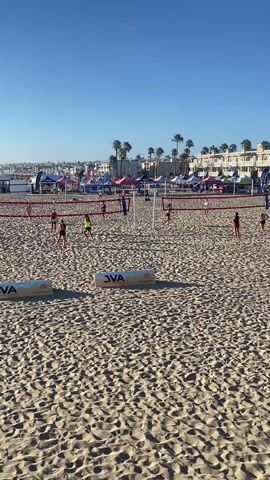 The video shows a beach volleyball court on a sunny day. The sand is a light brown color, and there are footprints and patterns from the sand. In the foreground, two white inflatable boundary markers with the JVA logo are visible. In the background, two beach volleyball nets are set up, with a few players on each side of the nets. The players are wearing athletic clothing, and some are in action, playing volleyball. Beyond the volleyball courts, there are rows of colorful tents and buildings, suggesting an event or a resort area. Palm trees are scattered among the buildings, and the sky is a clear, bright blue. The overall scene depicts a lively beach volleyball event.