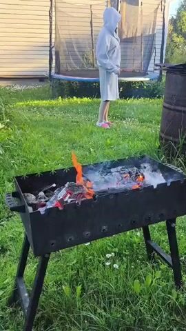 A person wearing a hooded jacket and shorts stands in a grassy yard in front of a metal grill filled with burning embers and flames. The person is facing the grill with their hands at their sides. A trampoline is visible in the background. The scene is lit by natural daylight.