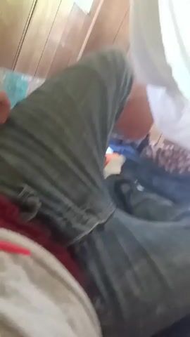 The video, shot from a first-person perspective, shows someone lying on a bed. The person is wearing gray jeans and the feet are visible. A white sheet hangs above the bed, and there is a blue blanket and a white piece of paper on the bed. The walls appear to be made of wood paneling. An air conditioning unit is visible near the ceiling.