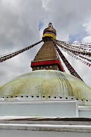 The video shows a close-up view of the Boudhanath Stupa in Kathmandu, Nepal. The stupa is a large white dome with a gilded spire, topped with prayer flags. The iconic "Buddha eyes" are visible on the side of the stupa. The sky is cloudy.