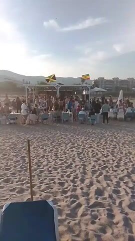 The video depicts a crowded beach scene with a white structure resembling a bar or stage at the center. Two Jamaican flags are visible flying from the structure. Numerous people are gathered, some standing and socializing, while others are seated at tables and chairs on the sand. The sand is a prominent feature of the foreground. The overall atmosphere suggests a lively outdoor event or gathering.