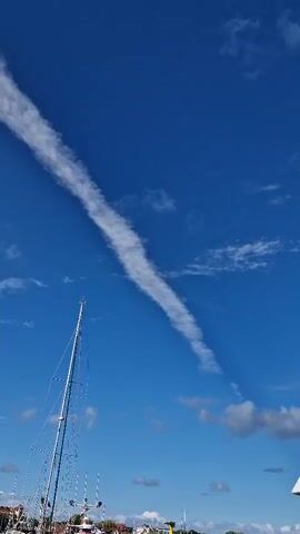 The video shows a clear blue sky with a long, thin cloud streaking across it. In the foreground, a tall mast of a sailboat is visible, with a few other boats and trees in the background. The sun is shining brightly, and there are a few smaller clouds scattered around. The camera pans slightly to reveal two streetlights on curved poles.