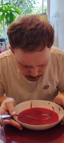 A man with red hair and a mustache is shown in the foreground, leaning over a bowl of red soup and holding a spoon. He is wearing a light-colored t-shirt. The background is blurred but appears to be an indoor setting with natural light coming from a window, suggesting a daytime scene. The man is looking down at the soup. The timestamp indicates the video is very short, starting at 00:00 and ending at 00:05.