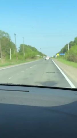 The video shows a car driving on a paved road. The road has two lanes going in each direction. The sky is blue. There are trees lining the road on both sides. There are other cars driving on the road. There are power poles along the side of the road. A road sign is also visible. The view is from inside the car, looking out through the windshield. The dashboard of the car is visible.