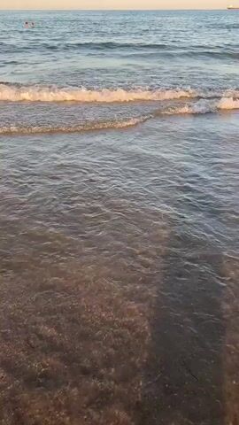This video shows a serene beach scene at Grao, Spain. The gentle waves of the ocean are rolling onto the sandy shore, creating a soothing sound and visual experience. The water is clear, allowing the sandy bottom to be visible, and it reflects the warm hues of the sky. The sun is setting, casting a golden glow over the water, indicating it is late afternoon or early evening. Two people are visible swimming in the distance, enjoying the calm sea. The overall atmosphere is peaceful and relaxing, suggesting a pleasant day at the beach. There are no visible objects or text.