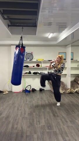 A person with blonde hair and tattoos on their arm is kicking a blue punching bag in a room. The person is wearing a black and white checkered top and dark pants. A second person, wearing a white t-shirt and green shorts, is in the background, also interacting with the punching bag. The room appears to be a gym or training space, with shelves in the background holding various items, including a dartboard and stuffed animals. A hula hoop is on the floor. The flooring is wood-like, and the ceiling is white.