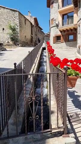 This is a static shot of a picturesque village street in Setcases, Spain. The scene is dominated by an old stone building on the left and a multi-story stone building with wooden shutters and a balcony on the right. A narrow channel of water flows down the center of the street, divided by a decorative metal fence. Red geraniums in a planter add a vibrant splash of color to the right side of the channel. The sun is shining brightly, indicating it is daytime, and the sky is a clear blue, suggesting fair weather. The architecture and the stone construction of the buildings are characteristic of a traditional mountain village. There are no people visible in the frame. The overall impression is one of tranquility and rustic charm.