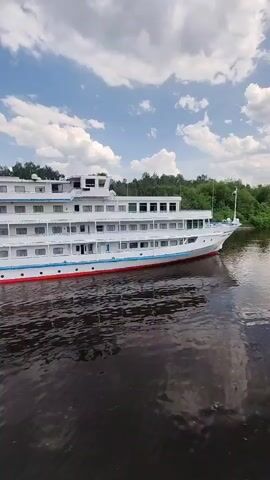 A cruise ship sits on a dark, glassy river with lush green trees lining the bank on the right.  The white ship has multiple levels and a red and blue stripe running along the hull. The sky is bright blue with fluffy white clouds. The ship’s reflection is visible in the water.
