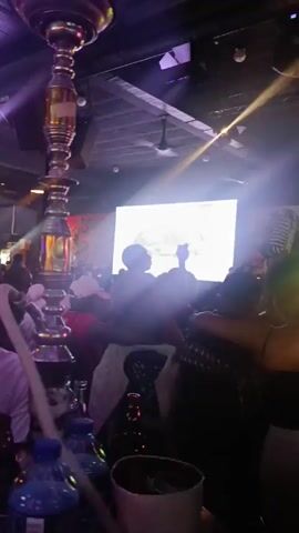 The video shows a crowded bar or nightclub with people dancing and enjoying the music. A live band or DJ is performing on a stage with a "Jameson Black Barrel" sign in the background. Colorful stage lights illuminate the space, and there are bubbles floating in the air. People are standing close to each other and moving to the music. The atmosphere appears energetic and lively.