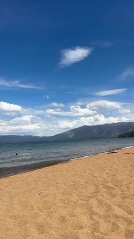 A video captures a sunny day at a sandy beach with calm, blue water and a mountain range in the distance under a bright blue sky with scattered white clouds. A gentle wave washes onto the shore. Several people are visible in the water, swimming and paddling on what appear to be small watercraft. The overall atmosphere is one of leisurely recreation.