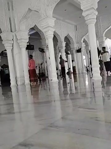 The video shows the interior of a large, white building with many tall, white columns and arches. The floor is made of a light-colored, reflective material, giving the appearance of a mirror. Several people are present, some walking, some standing, and one kneeling. The individuals are dressed in a variety of clothing styles. The overall atmosphere is bright and airy, reflecting the light from the windows and shiny floor.