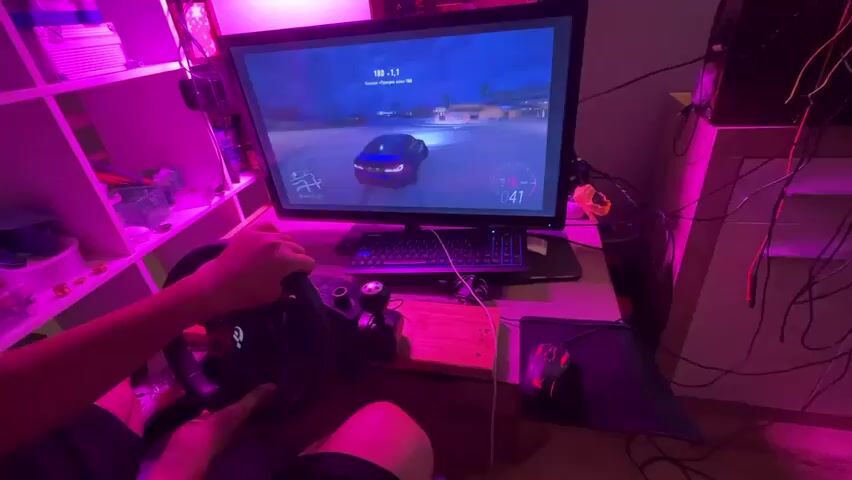 A first-person perspective video shows a person playing a racing video game called Forza Horizon 5 on a computer. The player is using a Logitech G920 racing wheel and pedals. The game is displayed on a large monitor, and the room is illuminated by pink LED lights. The player's hands are on the steering wheel and the gear shifter. The game shows a blue car driving through a street at night. The video is 4 seconds long.