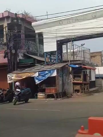 The video shows a street scene with buildings and vehicles. In the foreground, there are several motorcycles parked on the side of the road. A black car is parked on the left. Behind the motorcycles, there is a small shop with an awning. The building behind the shop has multiple levels and balconies with decorative railings. Signs are visible on the buildings, including one that says "CEPAT" and another with what appears to be a logo. Further down the street, a white SUV and a blue car are parked. Another building has signs for "GMW FOAM" and "NAPOLY BIGDREAM BIG FORM BIG." A yellow truck is parked on the left side of the street. The environment appears to be urban, with overhead electrical wires and poles. The overall impression is of a commercial street in a town or city.