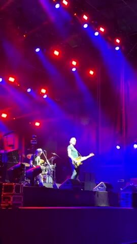 The video shows a concert performance with dramatic lighting. A man is playing a guitar on a stage, illuminated by red and blue spotlights. To his left, a drummer is seated behind a drum kit, also visible in the stage lights. The stage is elevated, and equipment such as speakers and other stage gear are present. The background is dark, with stage rigging and fabric visible. The overall atmosphere appears to be that of a live music event.
