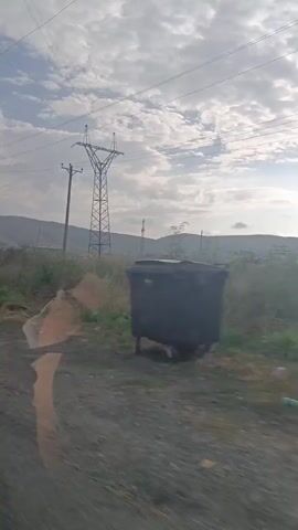 This is a video taken from a moving vehicle. The scene shows a rural landscape with rolling hills in the background under a cloudy sky. In the foreground, there are power lines supported by tall metal and wooden poles. Vegetation, including tall dry grass and some green bushes, lines the side of the road. A large metal pipe runs horizontally across the middle of the frame. The reflection of the vehicle's interior, specifically a mirror, is visible in the lower left of the frame. The camera is panning slightly, giving the impression of movement through the environment.