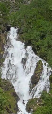 The video shows a tall, cascading waterfall with multiple tiers, surrounded by lush green vegetation. The water is white and foamy as it rushes down the rocky terrain. There are patches of moss and grass visible on the rocks adjacent to the waterfall. Towards the bottom of the falls, at least one person can be seen standing amidst the water. The overall environment suggests a natural, possibly mountainous, setting.