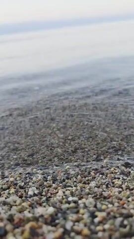 The video shows a close-up, low-angle view of a pebble beach with gentle waves lapping at the shore. The pebbles are small and varied in color, including shades of grey, white, brown, and pink. The water is clear and shallow, allowing the pebbles beneath the surface to be visible. The horizon is blurred in the background, with a light, overcast sky. The waves are small and create subtle ripples on the water's surface. The overall impression is one of tranquility and natural beauty.
