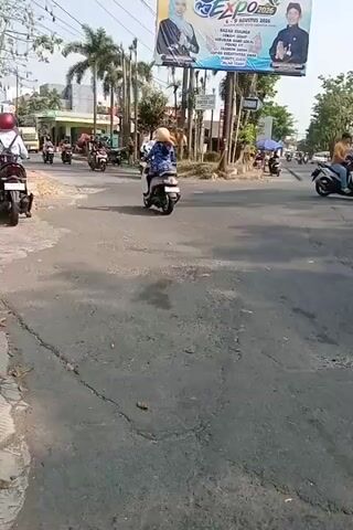 The video shows a busy intersection with several motorcycles in motion. In the foreground, the asphalt road is cracked and weathered, with a concrete curb on the left. In the background, a large billboard displays an advertisement for an "EXPO 2020" event, featuring images of two individuals. Palm trees and other foliage are visible, as are several buildings, including what appears to be a green-colored establishment on the left. A traffic light is also present. The sky is bright and clear, suggesting daytime. The overall scene depicts a typical urban or suburban environment with moderate traffic.