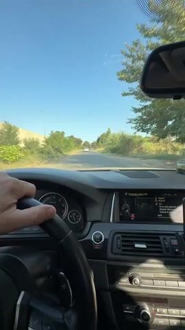 A first-person perspective video shows a hand on the steering wheel of a car driving down a paved road. The driver's side dashboard is visible, including the steering wheel, instrument cluster, and infotainment screen. Outside the car, green trees and foliage line both sides of the road. The sky is clear and blue. In the distance, several white cars are visible on the road. The video appears to be filmed during the daytime.