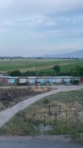 The video shows an outdoor scene with a row of garages. In the foreground, there are several rusted metal swing sets. A dirt road winds through a grassy area. In the middle ground, the row of garages, each with a differently colored door, runs horizontally across the frame. Behind the garages, there are fields of green crops stretching to the horizon. Mountains are visible in the distance under a cloudy sky. The overall atmosphere is rural and somewhat neglected. There are no people or animals visible in the video.
