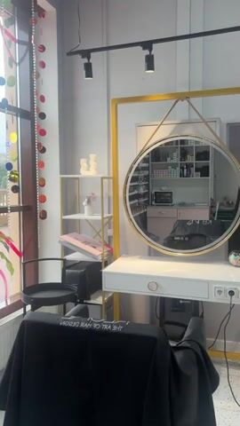 A young woman with long dark hair, wearing a black crop top and brown pants, takes a selfie in a round mirror in what appears to be a salon. The mirror is framed with gold and hangs from a leather strap. She is standing behind a white salon station with a gold frame. A black salon chair with a logo that says "Kano The Art of Hair Design" is in the foreground. In the reflection, shelves stocked with hair products, a microwave, a pink cabinet, a green sofa, and pink striped shelves are visible. The overall environment suggests a modern, well-equipped hair salon.