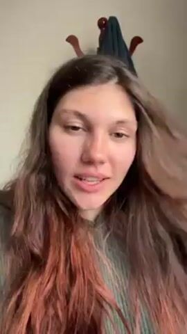 A young woman with long, brown hair that fades into a reddish-orange color at the ends is sitting in front of a white wall. The woman is wearing a green sweater and is running her fingers through her hair. Behind her, there is a wooden coat rack with a dark blue garment hanging on it. The woman is speaking, but the audio quality is poor and difficult to understand due to background music.
