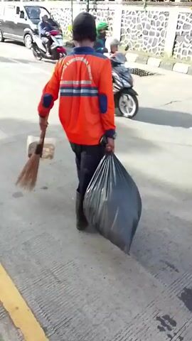 The video shows a street scene, likely in a Southeast Asian country. In the initial shot, a man in an orange jacket and black pants is seen sweeping the street and carrying a trash bag. The second shot shows a man in a green jacket walking on a curb. The street is busy with vehicular traffic including motorcycles and small green vans. A mountain is visible in the background. The sky is bright.