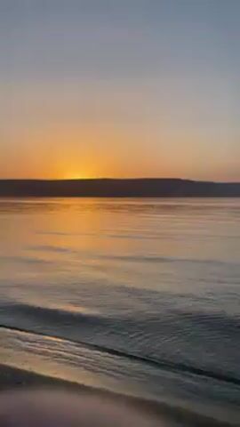 The video shows a woman fishing on a beach at sunset. The sun is setting over a body of water, with a strip of land visible in the distance. The woman is wearing a blue bikini top and black shorts. She is holding a fishing rod and reeling in a small fish. The water is calm and reflects the colors of the sunset. The beach is sandy with some small stones.