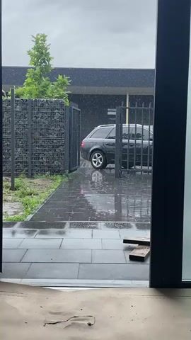 The video shows a view through a doorway onto a wet paved area outside. It is raining, with visible raindrops falling. A black Audi station wagon is parked behind a black metal gate. To the left of the car, there is a gabion wall filled with rocks and a green tree. The ground and paving stones are wet and reflective from the rain. In the foreground, two pieces of wood are lying on the paving stones. The sky appears overcast.