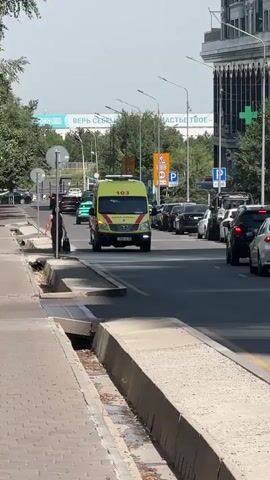 An ambulance with flashing blue lights is driving down a street. A woman in a black dress is standing on the sidewalk. Another person in a white shirt and black pants is walking near the ambulance. There are other cars on the road and trees lining the sidewalk.