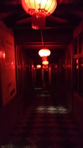 A dark hallway is illuminated by red lanterns. The lanterns are hung at intervals along the ceiling of the hallway, and their red light casts a dim glow on the walls and the checkered floor. The hallway appears to be empty.