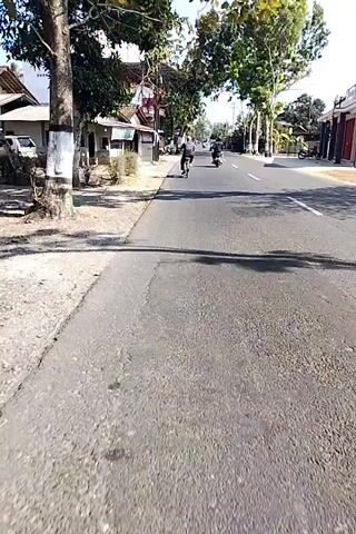 The video shows a first-person perspective of someone riding a bicycle on a paved road in a rural or suburban area. The road is lined with houses and trees on both sides. In the distance, other vehicles, including motorcycles, can be seen. The sun is shining, casting shadows on the road. The cyclist passes by small shops and houses. One shop has a banner with text and images on it. The cyclist's license plate is visible at the bottom of the screen.