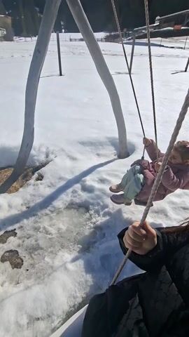 A woman is recording a video of herself and a young child on swings in a snowy environment. The swings are suspended from pale, tree-like wooden structures. The child, dressed in a pink jacket and patterned pants, is lying on the swing, smiling and holding onto the ropes. The woman, wearing a black jacket, is sitting on another swing, holding the ropes and looking into the camera with a smile. The ground is covered in snow, with some patches of bare earth visible near the base of the swing supports. In the background, there are buildings, trees, and a hillside. The scene is bright and sunny.