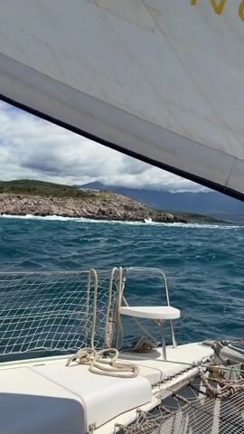 From the perspective of a sailboat, the video showcases a partly cloudy day on the water. The boat's white sail, partially visible at the top of the frame, is taut. A white deck chair and rope netting are visible in the foreground. In the distance, a rocky coastline meets the ocean. Mountains are partially obscured by clouds in the background. Waves break against the rocky shore. The water is deep blue.