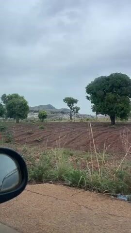 The video shows a view from a moving car, looking out at a rural landscape. The road is a light brown color and appears to be paved but possibly worn. The landscape consists of open fields of reddish-brown dirt that are lined in rows, possibly plowed for farming. Sparse green vegetation grows in patches across the fields. Several trees are scattered throughout the landscape, with most having full green canopies. In the distance, there is a rocky hill or small mountain, light in color and with sparse vegetation. The sky is overcast with a greyish tone. A power line with poles stretches across the field. The car's side mirror is visible in the lower left corner of the frame. The overall impression is a dull, cloudy day in a rural area.
