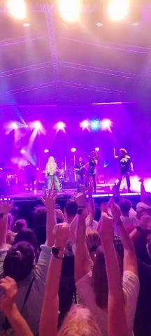 A group of five musicians, four women and one man, are performing on a brightly lit stage. The stage is bathed in purple and pink lights. The lead singer, a woman with long blonde hair wearing a colorful patterned dress, is in the center. To her right are two other women in dark, stylized outfits, and to her left are the male musician and another woman in a black outfit. The audience, a crowd of people with their hands raised in excitement, is visible in the foreground, below the stage. The overall atmosphere is energetic and festive, typical of a live concert.