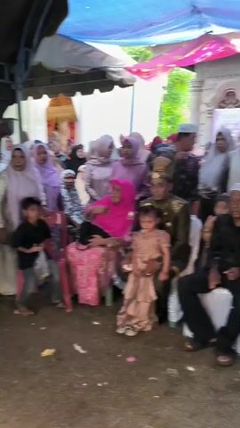 The video captures a gathering of people, likely a wedding ceremony, under a makeshift tent or canopy. The scene is crowded, with a mix of adults and children. Several women are wearing hijabs and traditional attire. In the foreground, a group of children in formal dresses are standing. Further back, an adult male is speaking into a microphone, while a woman in elaborate gold traditional wedding attire, likely the bride, is seated beside him, also in traditional dress and headwear. Other individuals are visible in the background, some seated and others standing, observing the proceedings. The ground appears to be dirt or packed earth, suggesting an outdoor or semi-outdoor setting. The overall atmosphere seems festive and ceremonial.