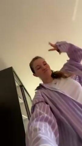 The video shows a woman filming herself while she's dancing indoors. She has her hair in a ponytail and is wearing a white t-shirt and a light purple striped shirt. In the background, there's a black cabinet and a painting with bright colors. She is moving the camera around as she dances. The room appears to be well-lit by natural light.