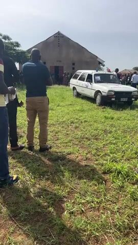 The video captures an outdoor scene in Panyam, Nigeria, during the daytime under a clear, sunny sky. In the foreground, a man in a dark blue shirt and khaki pants stands with his back to the camera. He is facing a white station wagon, which is parked on a grassy field. The car features a red cross symbol and a blue light on its roof, suggesting it is an ambulance or a medical service vehicle. Its license plate, "A2699BA," is visible. In the background, a stone or concrete building with a dark roof stands, likely a church or community hall, with people gathered near its entrance. Several individuals are also seen in the midground, near the white car, indicating a community gathering or event. Trees are visible in the far background.