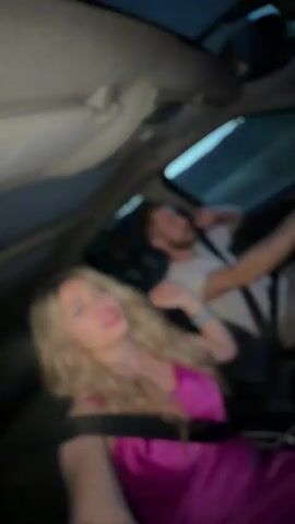 A blonde woman in a pink dress films a selfie video from the passenger seat of a car. A man drives the car, singing along to music and gesturing with his hand. They are both wearing seatbelts. It is dark outside.