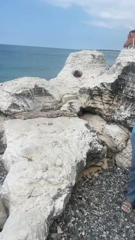 This media file captures a scenic view of the coast in Tsandrypsh, Abkhazia. The scene is dominated by large, weathered white rock formations leading down to a pebble beach and the turquoise waters of the sea. The sky is clear with a few scattered clouds, suggesting a pleasant, likely daytime weather condition.

In the foreground, the rough texture of the white rocks is evident, with some showing darker, possibly wet or mossy patches. A scattering of dark pebbles covers the beach area between the rocks. The sea stretches out to the horizon, appearing calm and vast.

Two individuals are present in the scene. On the right, a person is partially visible from the waist down, wearing a long, dark red skirt with a high slit, white sneakers, and white socks. They are standing on the rocks, looking out towards the sea. A leopard-print crossbody bag is visible. To the left and further back, a man in a white t-shirt and a dark baseball cap stands on the rocks, also facing the sea. Their presence suggests people enjoying the coastal scenery.

The overall mood appears peaceful and relaxed, with the focus on the natural beauty of the coastline. There is no visible text in the media file. The time of day is likely daytime, given the bright lighting and clear sky.