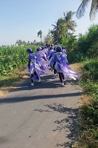 A group of people in matching purple outfits are walking down a paved road. One person is riding a motorcycle and appears to be following them. The road is lined with greenery and trees, suggesting a rural or outdoor setting. The sky is clear and blue.