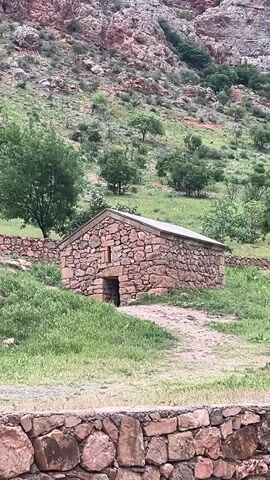 The video opens with an exterior shot of a small, rectangular stone building situated on a grassy slope, with a larger, tree-covered mountain in the background. A low stone wall is in the immediate foreground, and a dirt path leads up to the building's front entrance, which has a small, arched opening and a single, rectangular window above it. The scene transitions to a group of eight people, including one child, walking on a paved path alongside a larger, multi-story stone structure. This building exhibits intricate stone carvings, including numerous crosses, and features an exterior stone staircase. The people are dressed in casual clothing, with some carrying small bags or bottles. They are walking in a relaxed manner, and some are conversing.