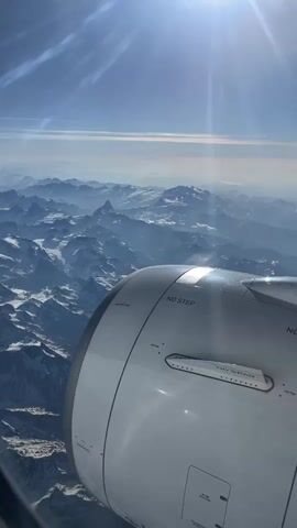 The view from an airplane window shows a snow-covered mountain range under a clear blue sky with the sun shining brightly. Part of the airplane engine is visible in the foreground, with the words "NO STEP" and "NO GRAB" printed on it. The mountains stretch out into the distance, fading into a hazy horizon.