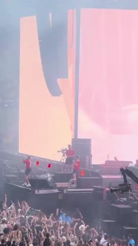 This is a video taken from a concert, likely at an indoor arena or stadium, given the large screens and vast audience. The stage is elaborately lit with bright spotlights and smoke effects. Several performers in red shirts are visible on the stage, some appearing to be singing or interacting. A camera rig is also present on the stage, suggesting professional filming. Below the stage, a large crowd of concertgoers can be seen, many holding up their phones to record the event. The atmosphere appears energetic and exciting, with the audience actively engaged.

The video displays large screens showing a live feed of the concert, with images of a stadium packed with people. Text is visible on the screens, including "DUSSELDORF - EST." This indicates the event is taking place in Dusseldorf, Germany. The time of day is likely evening or night, as suggested by the concert lighting. The emotion conveyed is one of excitement and collective enjoyment of the music and performance.