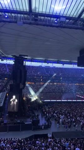 A wide-angle shot from above a stadium concert shows a massive crowd of people holding up their cell phones, their lights creating a shimmering effect. The stage is set with elaborate lighting and scaffolding, and a performer is visible on stage, bathed in spotlight. A large screen on the left side of the stage displays a black and white image of a man. The stadium ceiling is illuminated with blue and white lights, giving the scene an ethereal glow. Smoke or fog machines are being used on stage, adding to the dramatic atmosphere. The overall impression is one of a large-scale, high-energy musical event.