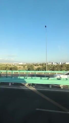 The video begins with a view from a moving vehicle, looking out the window at a bright blue sky and a distant cityscape. A green highway barrier runs parallel to the road, with a single lamppost visible on the left side. Trees and vegetation line the other side of the highway, leading towards the city skyline. The camera appears to be panning slightly to the right as the vehicle moves forward. The narration in the video states "And that's it. We're leaving China. It's so beautiful here." The video captures a scenic view of a transition away from an urban area, emphasizing the beauty of the landscape.